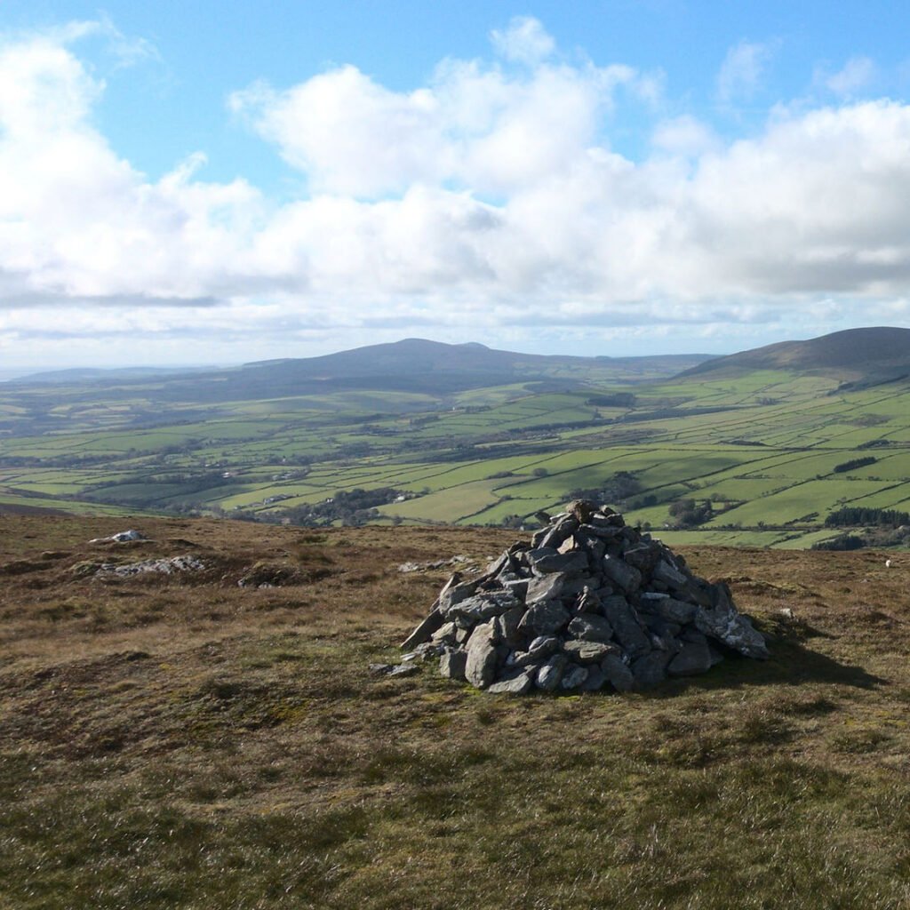 A view from the top of a moorland hill, with a pile of stones in the foreground. Rolling green smaller hills and valleys can be seen below and a taller hill with another nestled to it's right are seen in the far distance. The sky is blue with large fluffy white clouds.