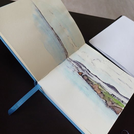 An open sketchbook showing an ink and watercolour sketch of a headland and the sea.