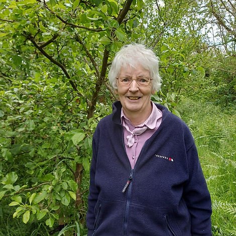 A photo of an older lady standing in front of trees and vegetation. She is smiling at the camera.