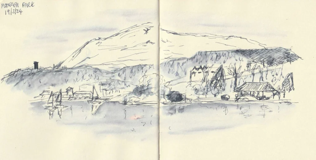 A sketch of Mooragh Park in Ramsey in grey watersoluble ink. The sketch shows small boats on a lake with trees and bushes, some buildings and then hills, behind.