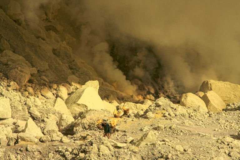 A man carries baskets full of bright yellow rocks amid a landscape of paler yellow rocks and yellow vapours.