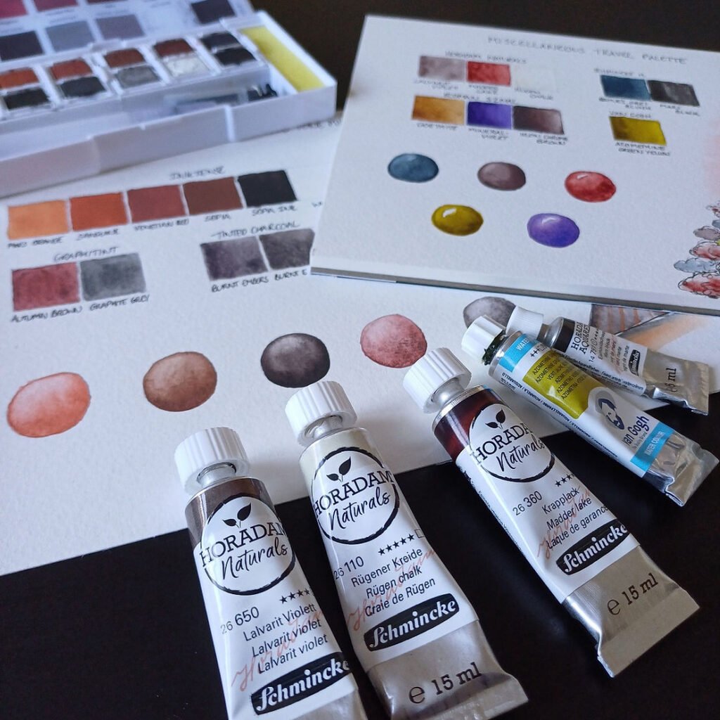 A photo of two swatch sheets with watercolour and other waterbased media swatched in mainly browns, oranges and greys. There is a purple, blue and greenish yellow colour too. In the foreground are five tubes of paint and in the background a white palette containing pans of paint.