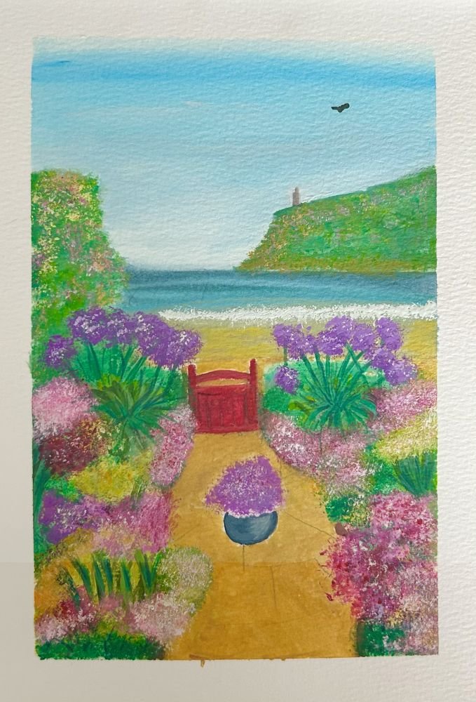 A colourful garden, filled with flowers with a red garden gate. The sea and a beach is visible just beyond the garden with a headland in the distance. The headland has a stone tower on it.