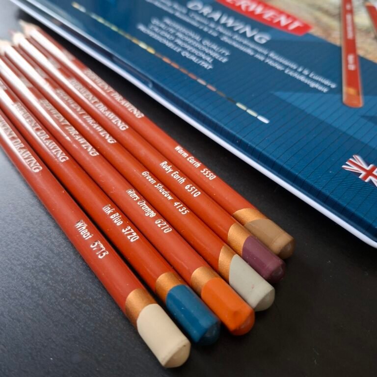 20251017_091446(1) Six coloured pencils from the Derwent Drawing range in 'Wheat', 'Ink Blue, 'Mars Orange', 'Green Shadow', 'Ruby Earth and 'Warm Earth' colours. They are on a black table in front of a pencil tin.',