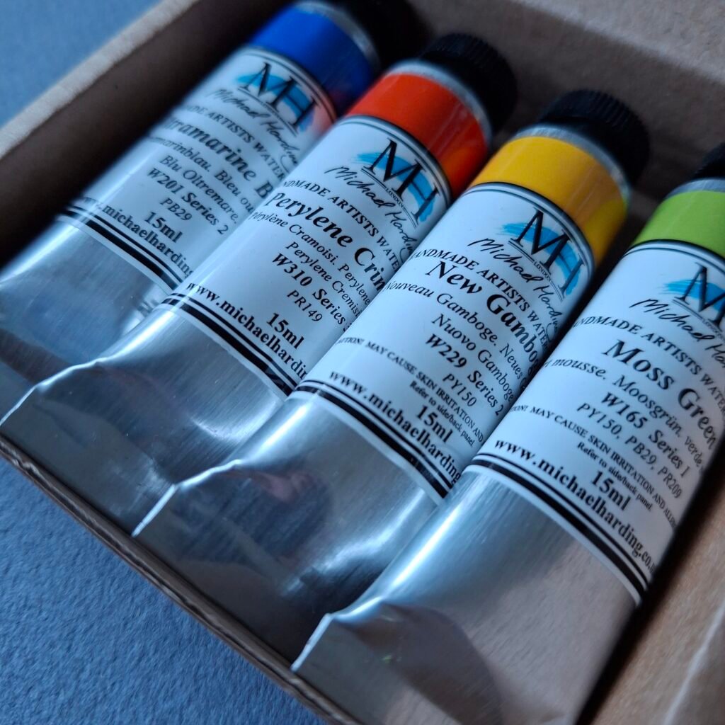 A set of four tubes of watercolour paint in a small cardboard box. The colours are a blue, red, yellow and a green.