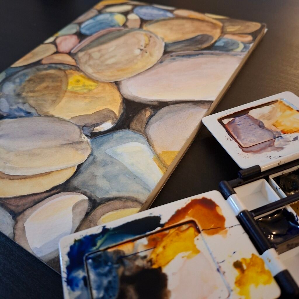 Watercolour painting of pebbles in a block of paper next to an open palette showing half pans of paint and used mixing areas. They are resting on a black table.