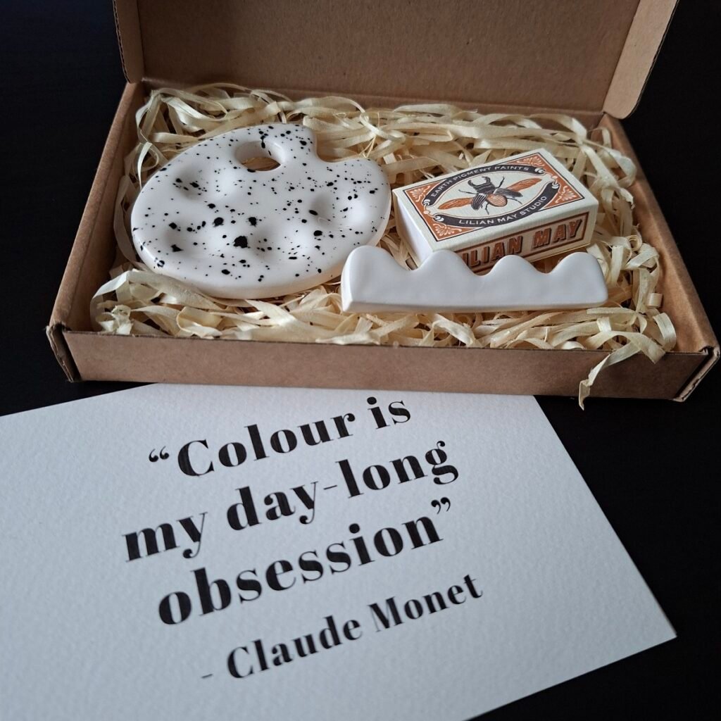 An open brown box containing a matchbox printed with a moth design and an artist palette shaped small ceramic palette. There is also a white ceramic brush rest in the box on top of the straw-like paper packaging material.