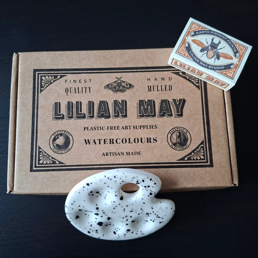 A brown box with a graphic reading Lilian May on the front with a matchbox printed with a moth design and an artist palette shaped small ceramic palette.