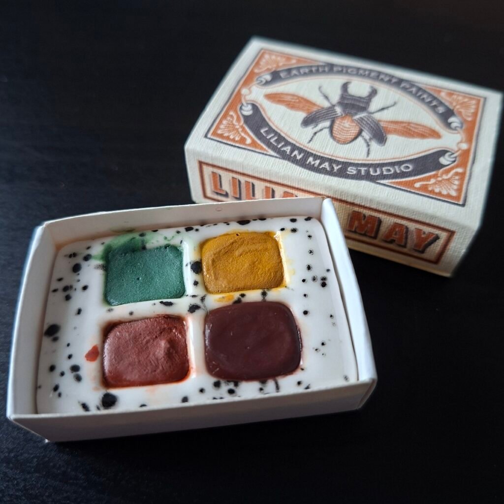 An open matchbox printed with a moth design showing the small ceramic palette of four paints within.
