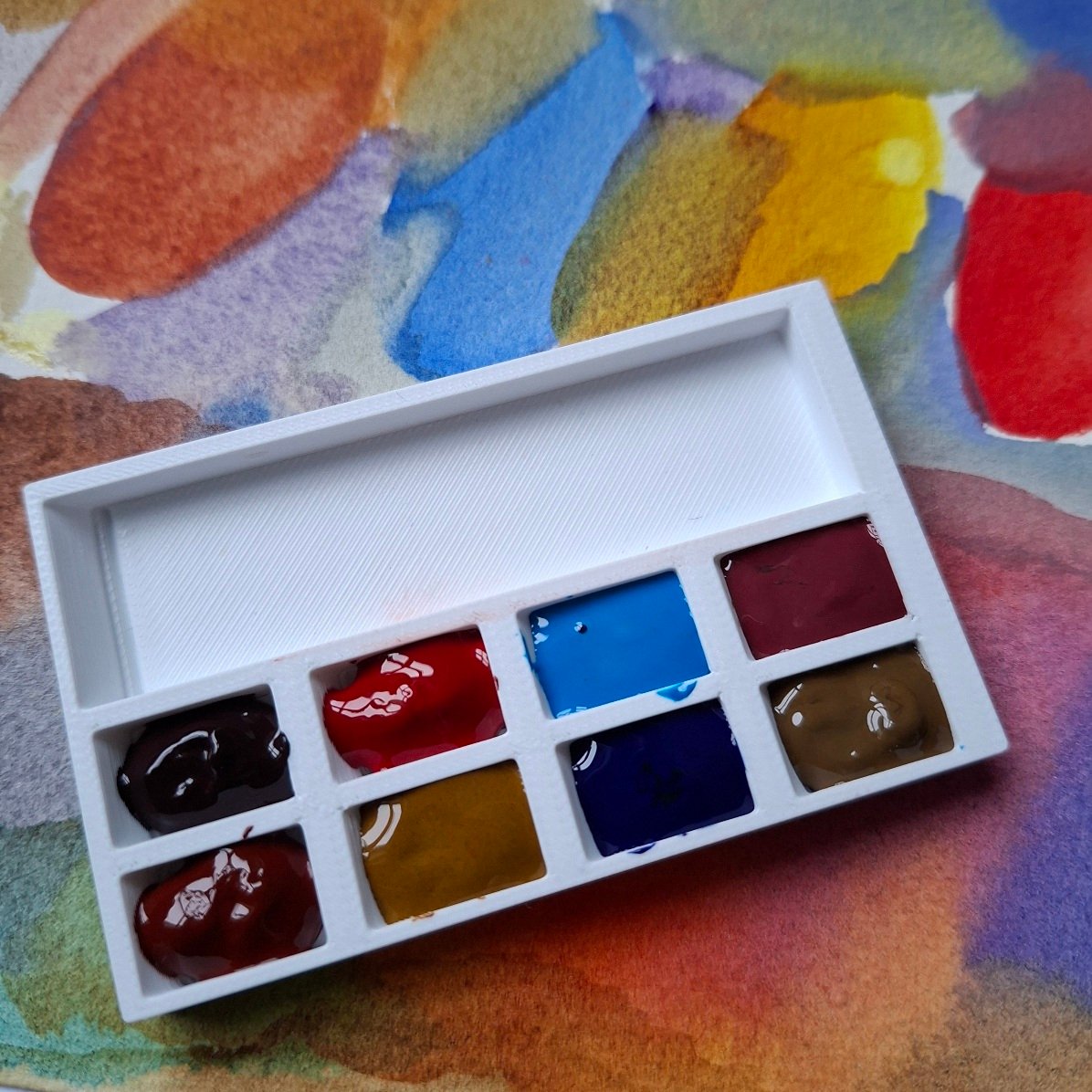 A small white 3d printed plastic palette filled with watercolour paint in its 8 wells. The palette sits on a piece of watercolour paper that's been painted with the colours of paint in spots that have run into each other.
