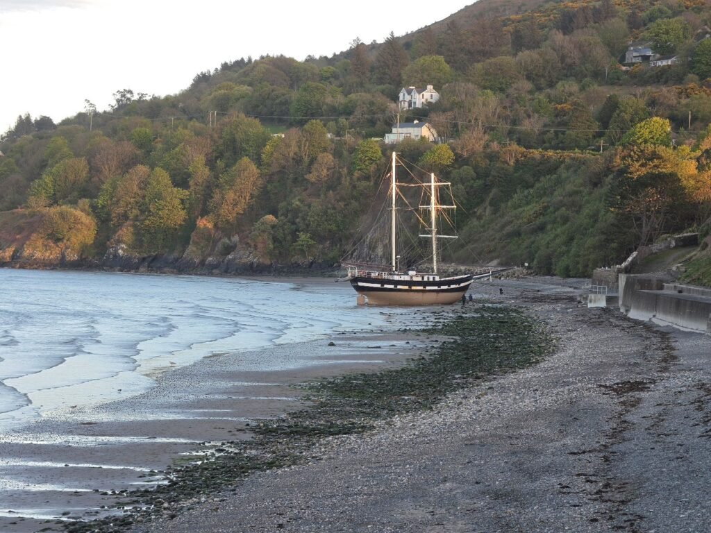 Photo of a beached, two masted tall ship on a sandy shoreline with green cliffs behind.